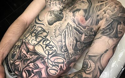 Arm und Bauch bedeckt mit Chicano Tattoos, betende Hände, Jungfrau Maria, Kreuz, Frau, Lettering